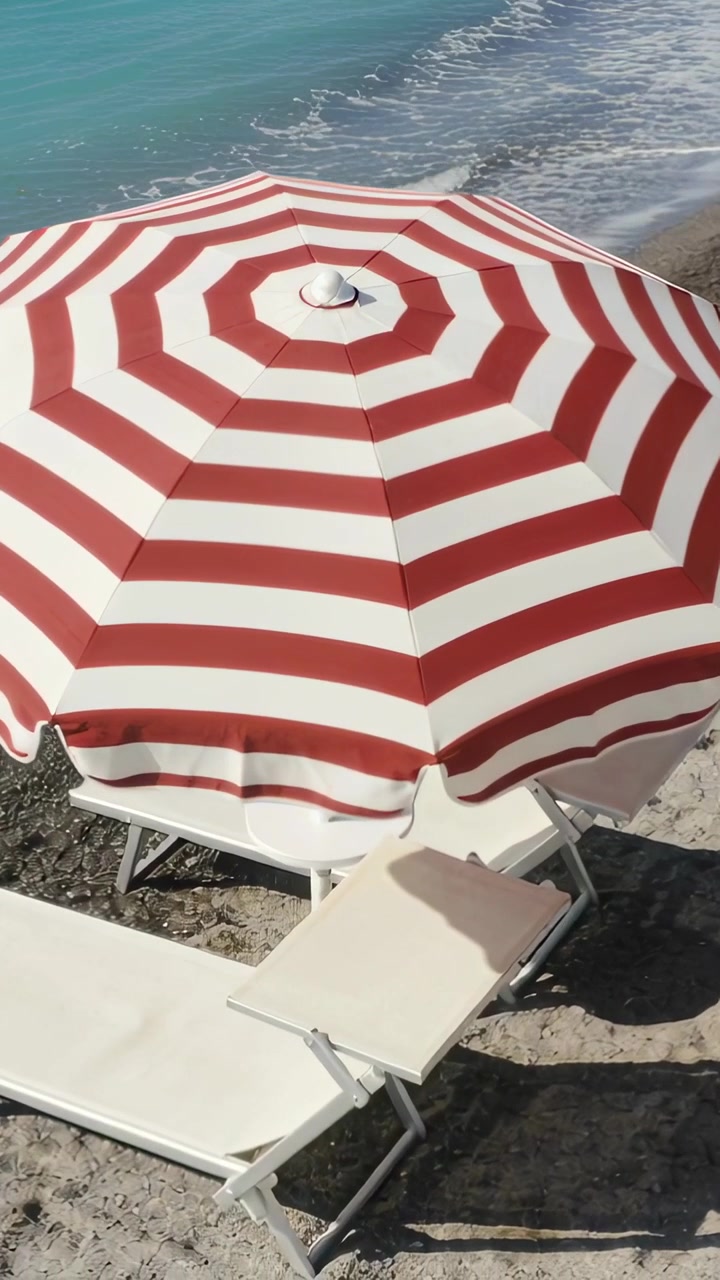 Video verticale: Telecamera che panoramica un ombrellone a strisce mentre le onde lambiscono la riva di ciottoli e le sedie si spostano.
