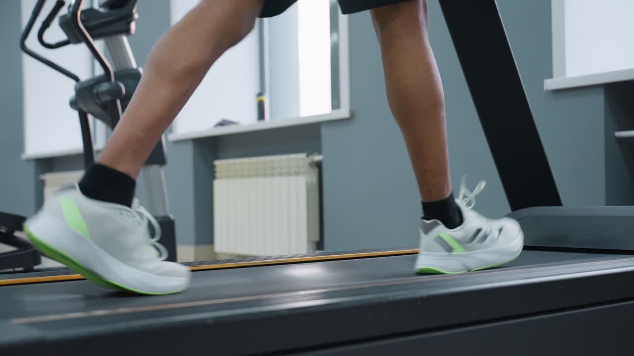 lower angle side view of person in black shorts and white sneakers powering treadmill workout in gym, focus on calf muscles and dynamic movement, blurred elliptical machine and radiator in background