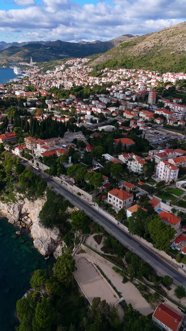 Aerial portrait cinematic drone soars over Dubrovnik town, capturing terracotta rooftops and winding streets with majestic mountains in the background under a clear, vibrant blue sky overhanging