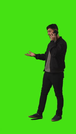 Vertical-Video-Full-Length-Shot-Of-Young-Man-Talking-On-Mobile-Phone-Walking-Into-Frame-Standing-Against-Green-Screen