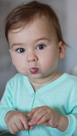 adorable child in blue shirt sitting at feeding table with his mouth full. kid with spoon in hands stretching his hand to the camera. vertical video