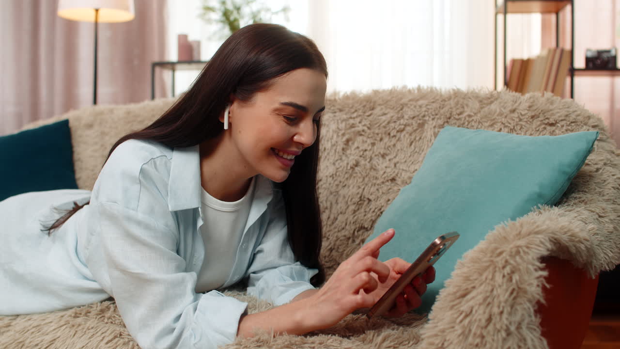 Woman lying on sofa wearing headphones scrolling social media feed on mobile phone relaxing calmly