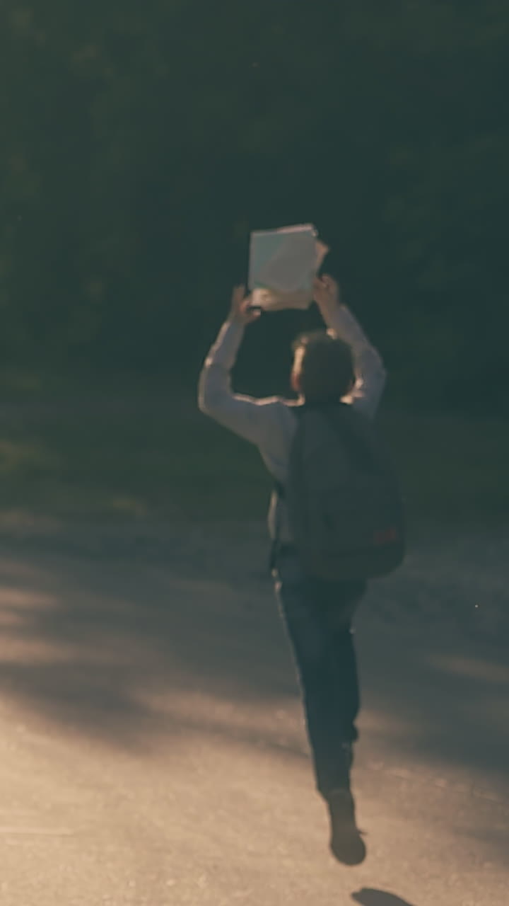 young pupil in denim jeans and shirt runs along asphalt road and throws up white sheets slow motion backside view