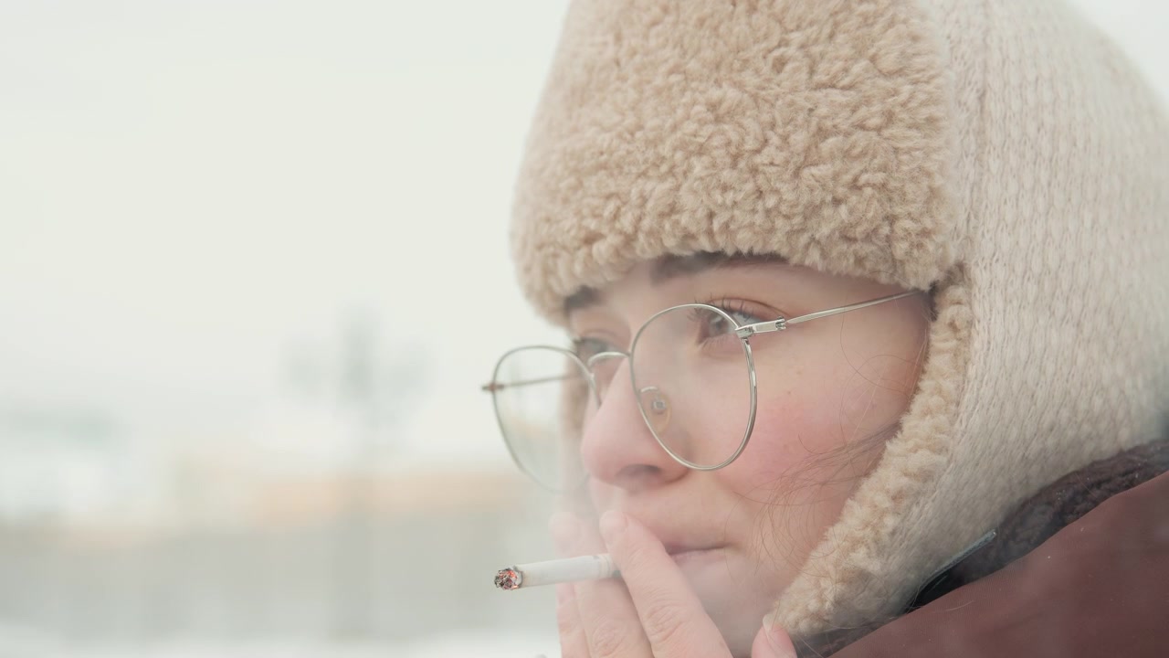 Primo piano di testa e spalle di una giovane donna che indossa un cappello con paraorecchie foderato di pelliccia e occhiali rotondi all'aperto con tempo nevoso e coperto, respiro visibile ed espressione calma e pensierosa, con donna, femminile.