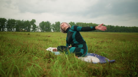 middle-aged woman with grey hair seated on yoga mat practicing side bend pose, hands stretched over her head, in a vast grassy field under cloudy skies, with trees lining the distance