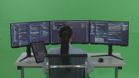vista trasera de una desarrolladora asiática trabajando con una tableta mientras escribe código con múltiples pantallas de computadora en un estudio de pantalla verde