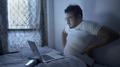 exhausted young man using laptop, working full night on bed and feeling pain in waist