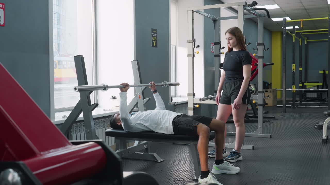 young girlfriend watches boyfriend perform weighted barbell bench press on flat bench inside bright gym with squat rack free weights and mirror in background while other member walks past