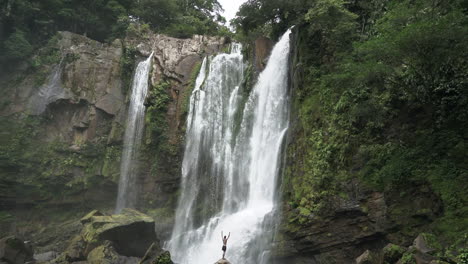 man stands with arms raised in joy in front of large rain forest waterfall