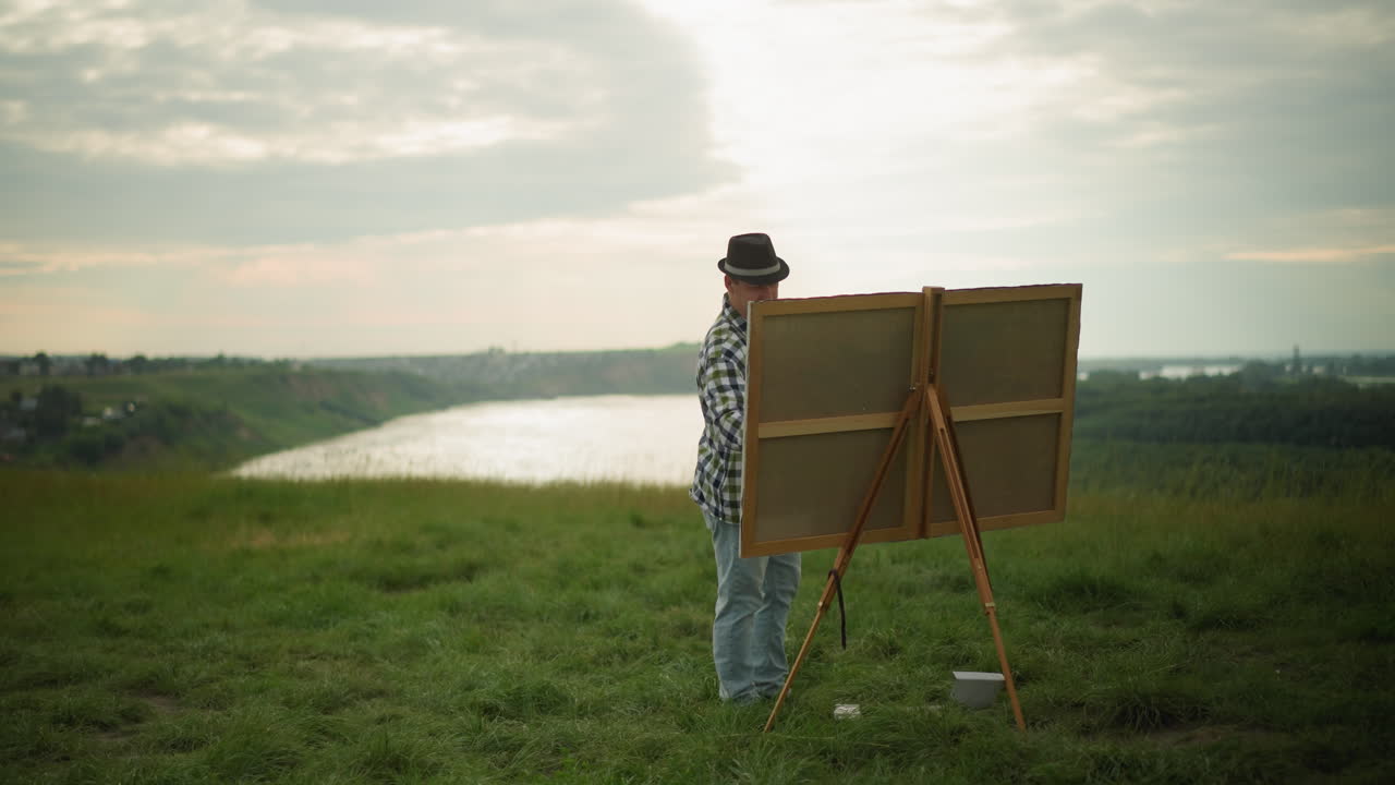 A sketcher in a black hat, checkered shirt, and jeans is engrossed in painting on a large canvas set up in a peaceful grass field by a serene lake. The calm setting, surrounded by natural beauty
