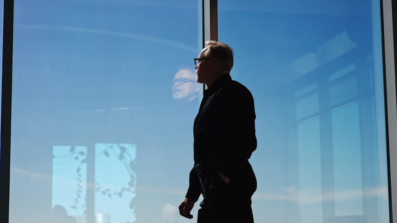 Confident pensive man wearing black shirt and trousers stands at the panoramic window. Man thinking over the issues looking at window. Low angle view.