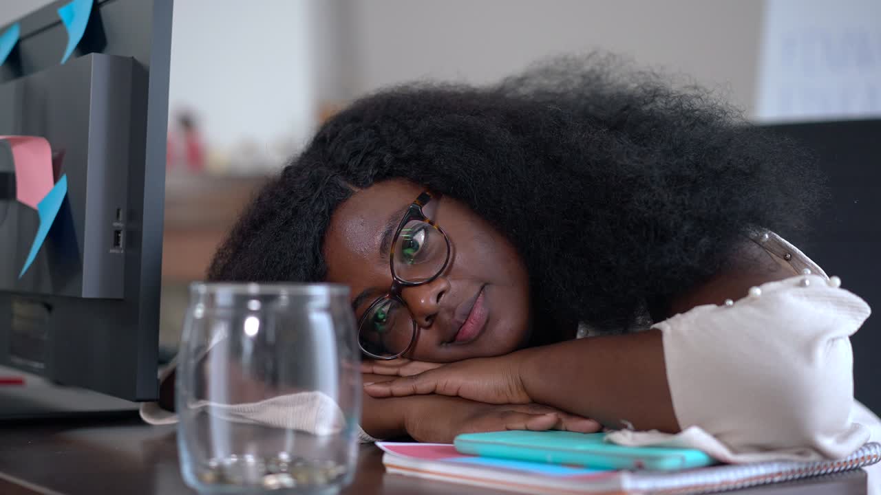 Close-up of sad African American young woman lying down on table looking at jar with coins in slow motion. Unsuccessful female freelancer indoors in home office thinking on revenue and expenses.