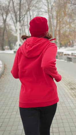 back view of woman jogging along snowy park pathway in red hoodie and black leggings beside benches bushes iron railing lamp posts white building distant cross monument conveying cold fitness motion