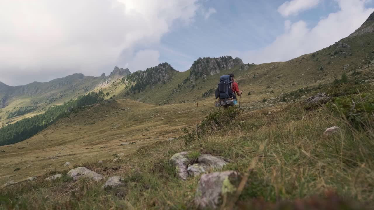 plano amplio en ángulo bajo de un excursionista masculino caminando solo por un hermoso sendero rural rodeado de montañas cubiertas de maleza en verano