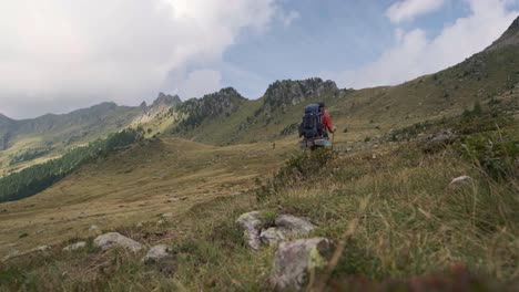 Low-angle-wide-shot-of-male-hiker-hiking-alone-on-beautiful-rural-trail-surrounded-by-overgrown-mountains-in-summer