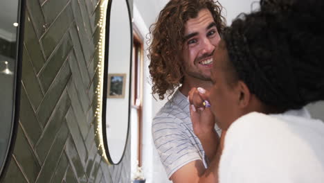 Diverse-couple:-a-young-African-American-woman-applies-makeup-to-a-young-Caucasian-man-at-home