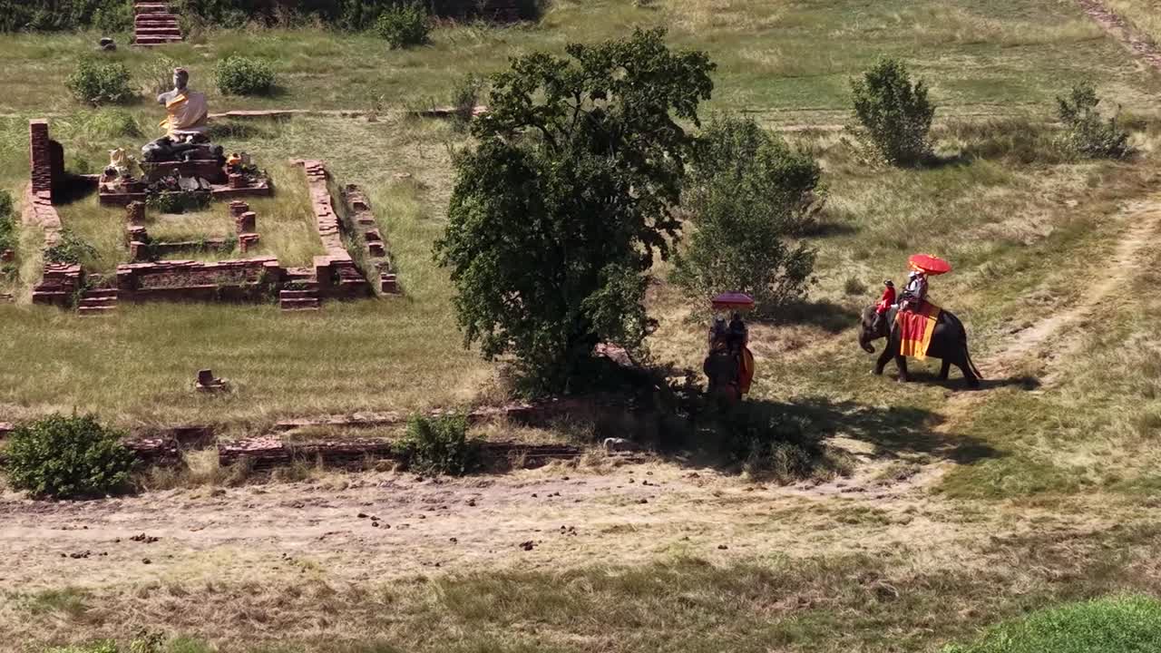 Drohnenaufnahme von Elefanten aus hohem Winkel, die TouristInnen durch einen historischen archäologischen Park in Thailand tragen
