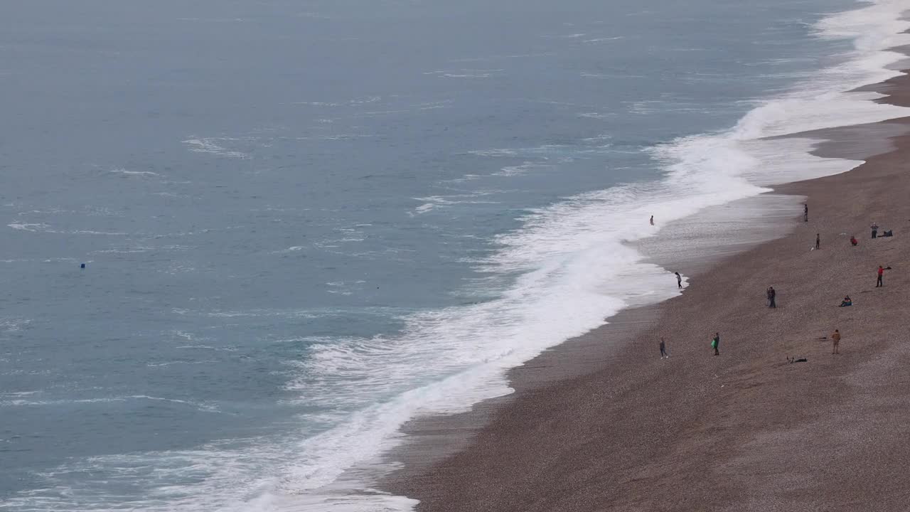 Wide coastal view with waves crashing on pebbled shoreline, scattered walkers moving along beach edge, white surf forming textured patterns, and calm overcast light covering Lara Beach in Antalya