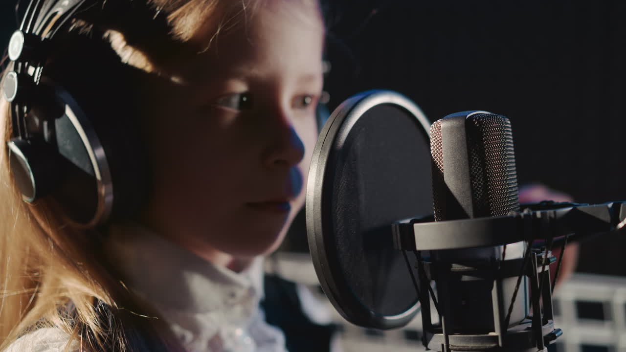 Child taps on microphone in recording studio. Curious little girl with headphones checkups mic by finger in sound insulated room. Vocal lesson