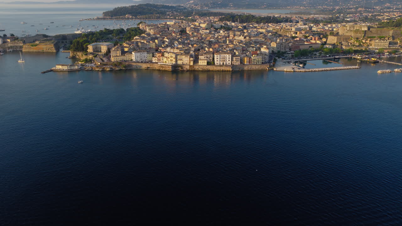 Swallows flying above rooftops of Corfu Old Town in soft blue light at sunrise, drone tilt up from ocean to cityscape