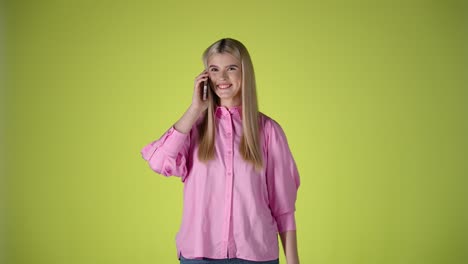 Una-Joven-Rubia-Caucásica-Habla-Alegremente-En-Un-Teléfono-Celular-Agitando-Sus-Manos,-Fondo-Cromático-De-Estudio,-Foto-De-Modelo-Infinito-Amarillo