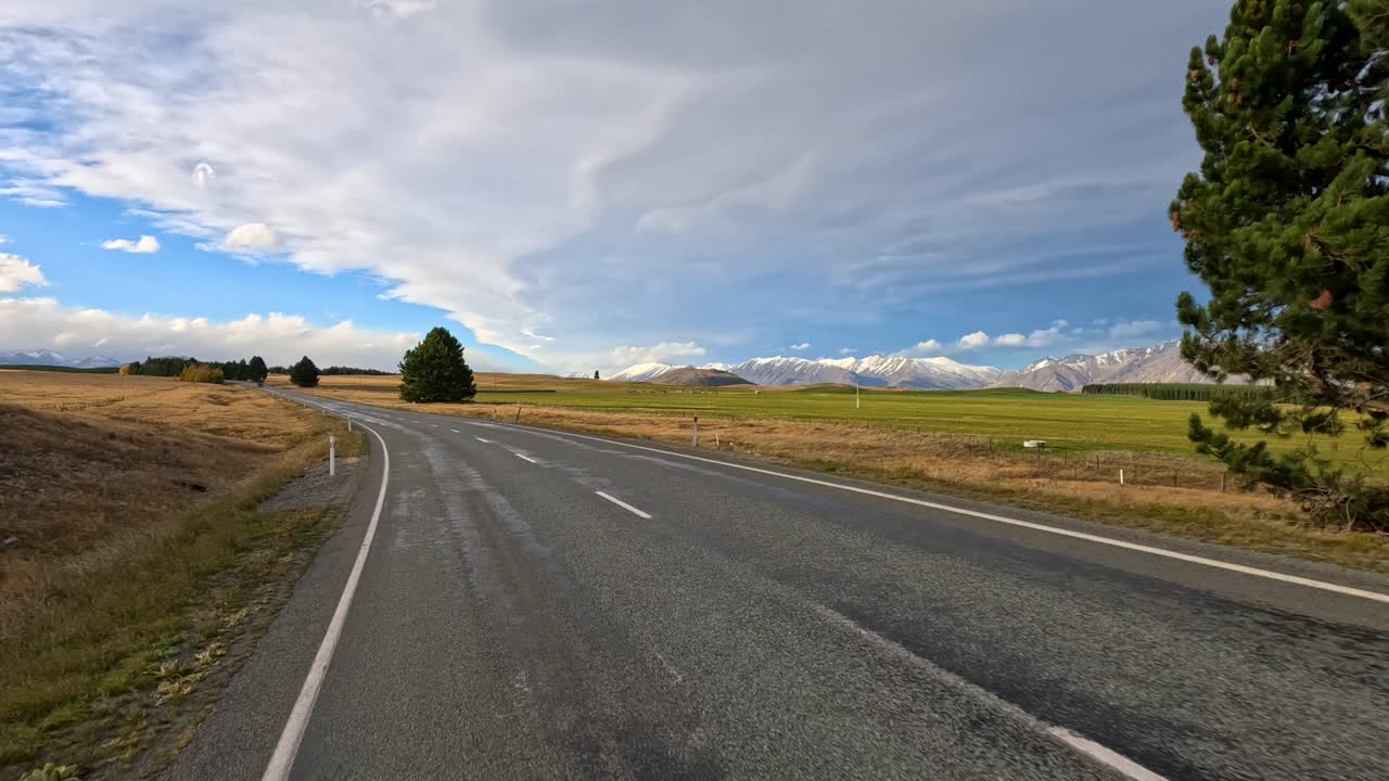 Un veicolo percorre una strada rurale oltre un albero solitario, con campi autunnali, nuvole drammatiche e montagne innevate in lontananza sotto la luce naturale del giorno.