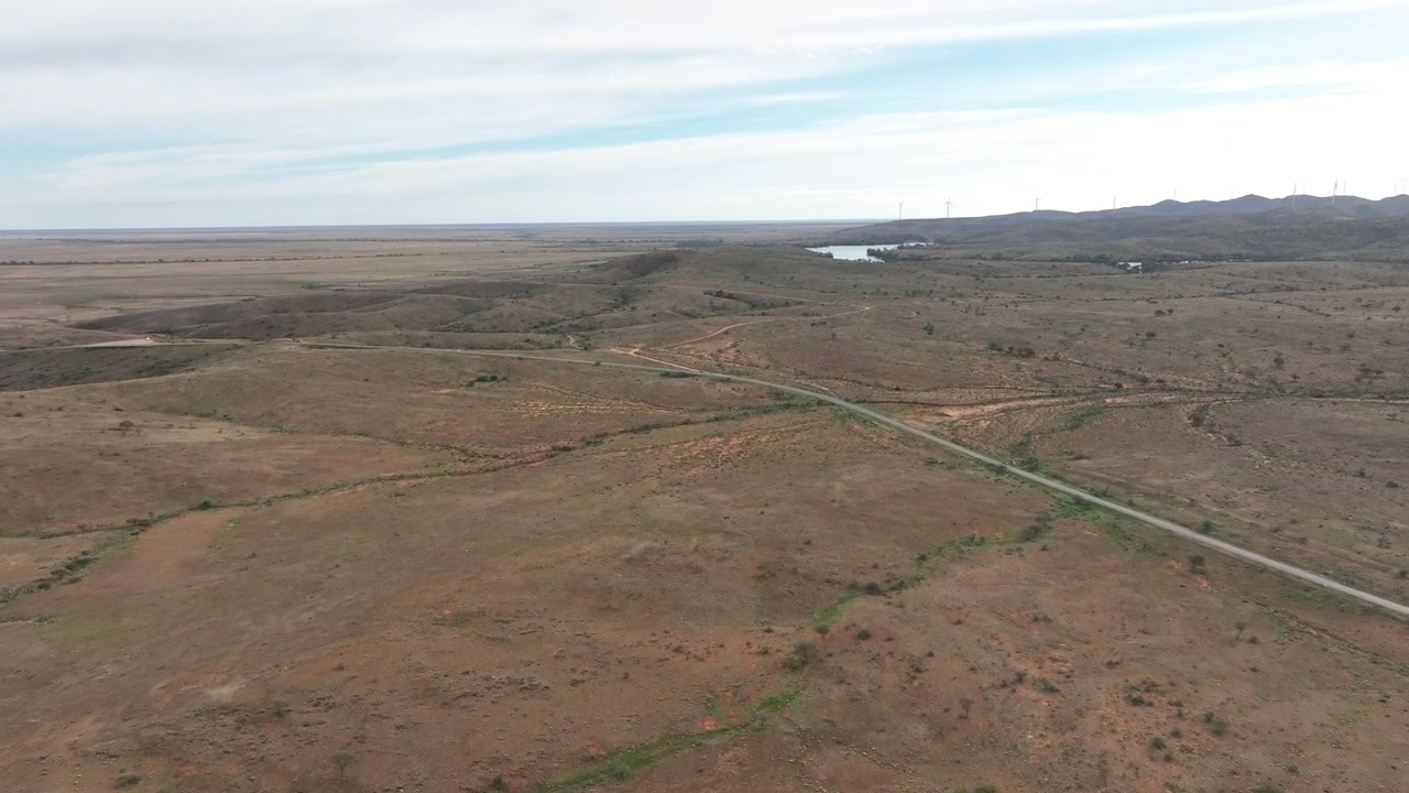 Rozsáhlý letecký pohled dronem přes odlehlé polosuché pláně a zvlněný terén poblíž Silvertonu a Broken Hill, s větrnými turbínami viditelnými podél vzdáleného horizontu.