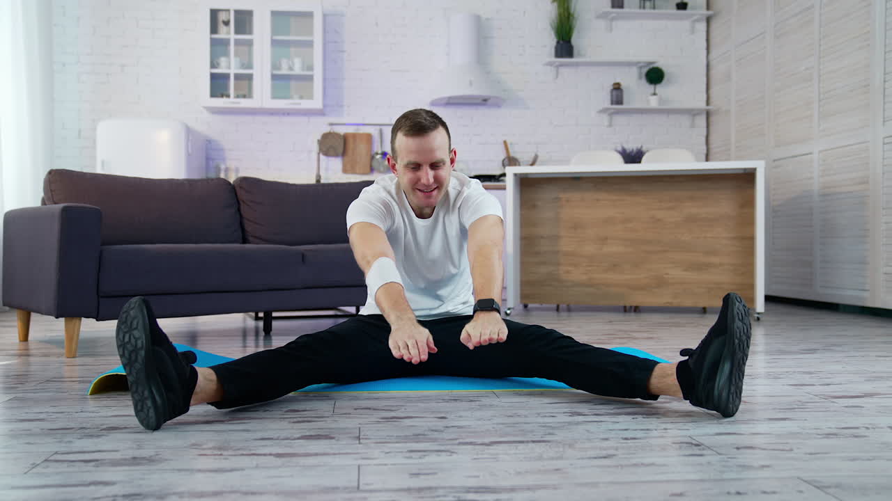 Young man doing workout at home. Guy is sitting on a mat and trying to do exercising during a quarantine period. Difficulties of doing workout at home.