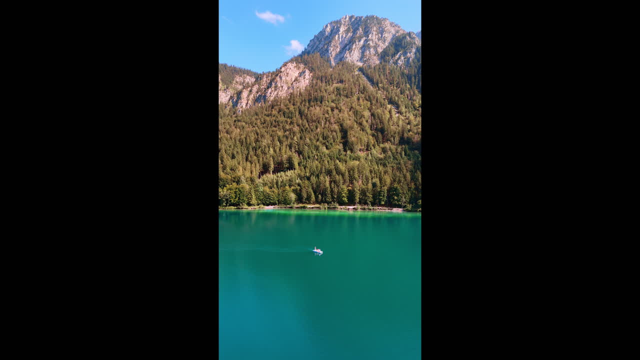 Footage over the turquoise lake with boats on the waterscape. Wooded rocks at backdrop. Bavaria, Germany. Vertical video