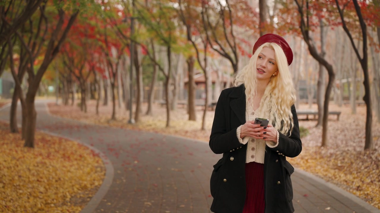 Blonde europäische Frau tippt auf ihrem Handy, während sie auf einem von Herbstblättern umgebenen Weg geht, Zeitlupe, sanftes Lächeln voller Staunen und Bewunderung.