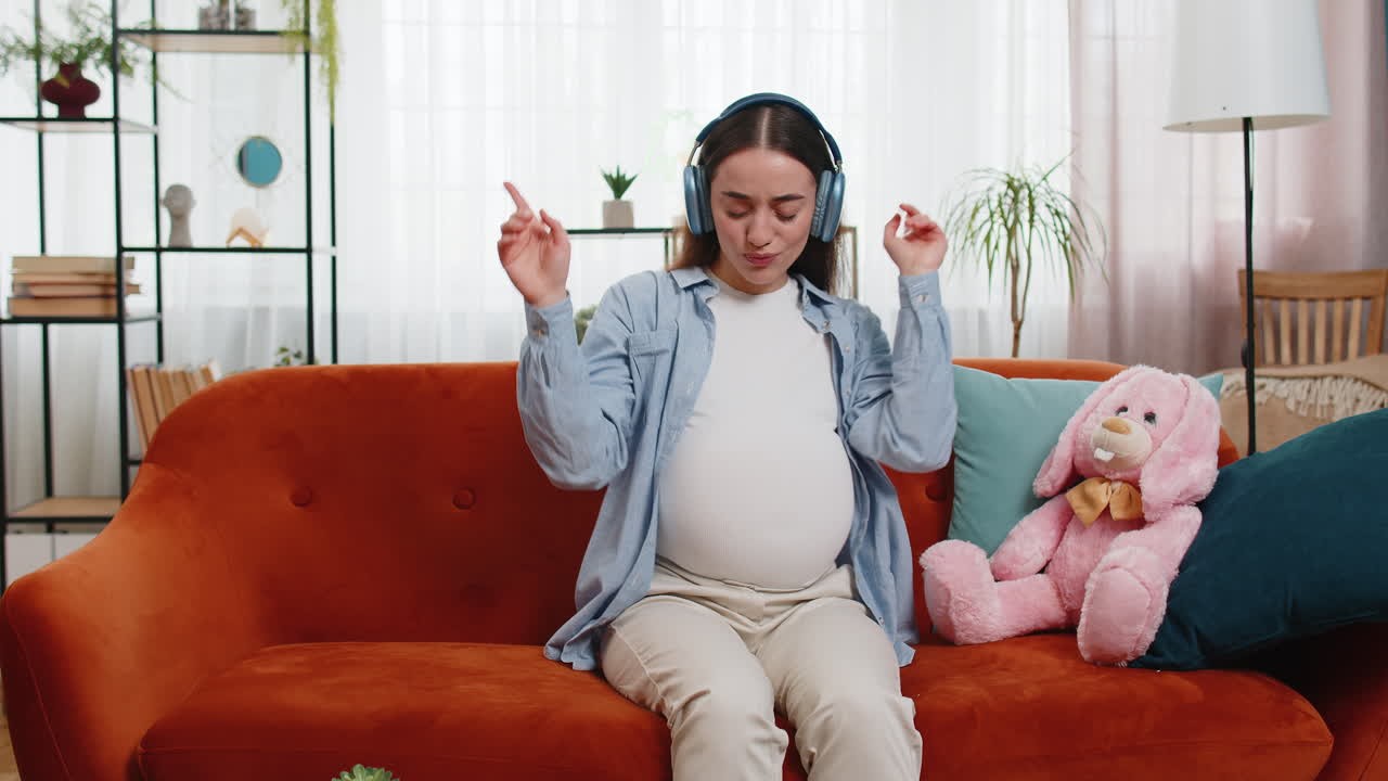 무선 헤드폰을 착용한 행복한 임신한 여성이 음악을 듣고 춤을 추고 휴식을 취하고 소파에서 축하하고 있습니다.