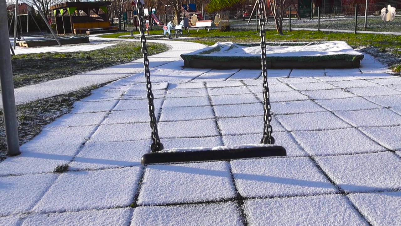 Empty swing with chains moving back and forth over a snow covered ground in a desolate children's playground on a sunny day, creating a melancholic and nostalgic atmosphere