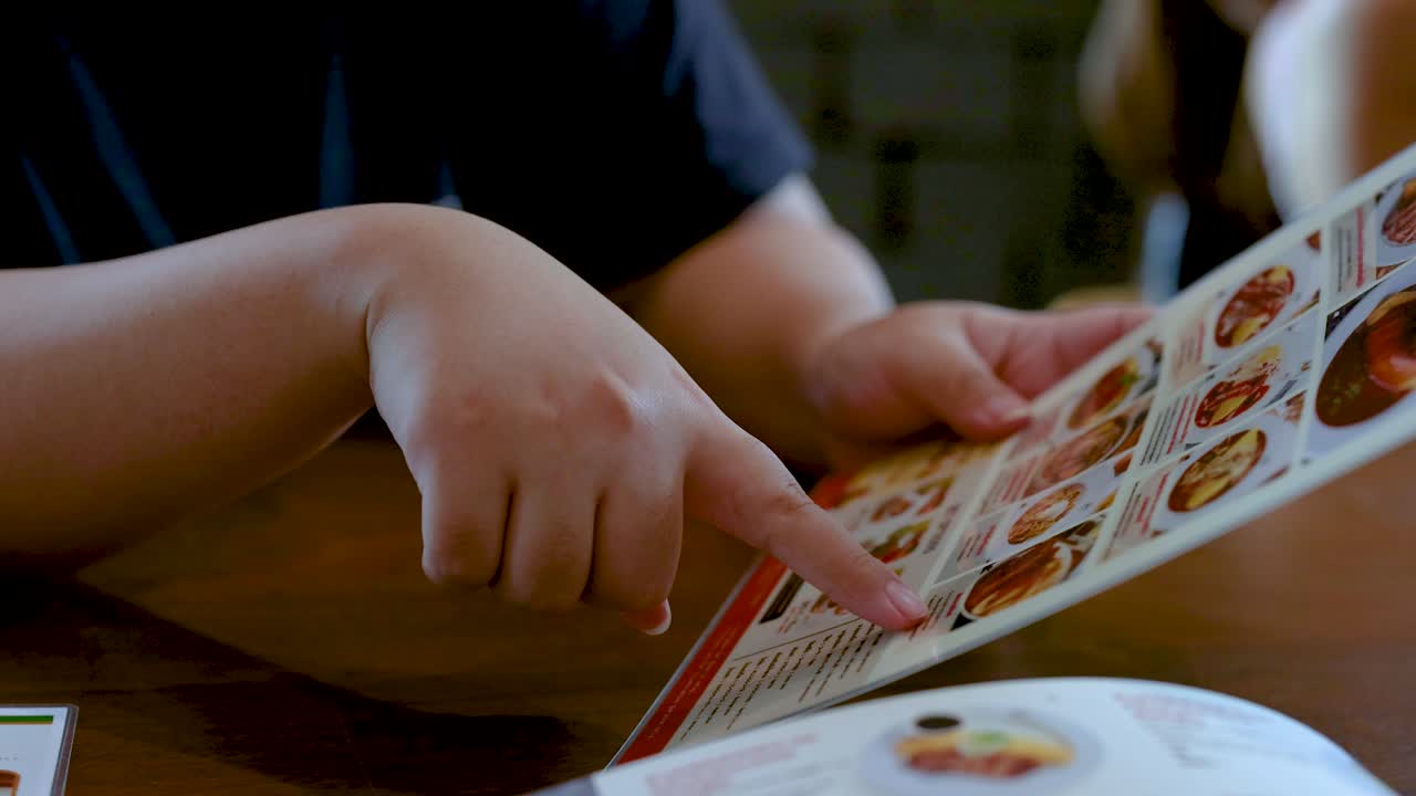 Person points at illustrated menu, selecting dishes at dining table in softly lit Bangkok restaurant