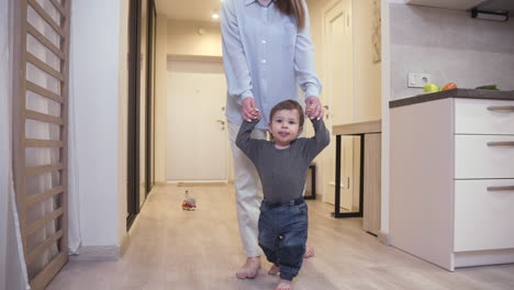 mother helping her baby to take his first steps from the corridor to the living room at home