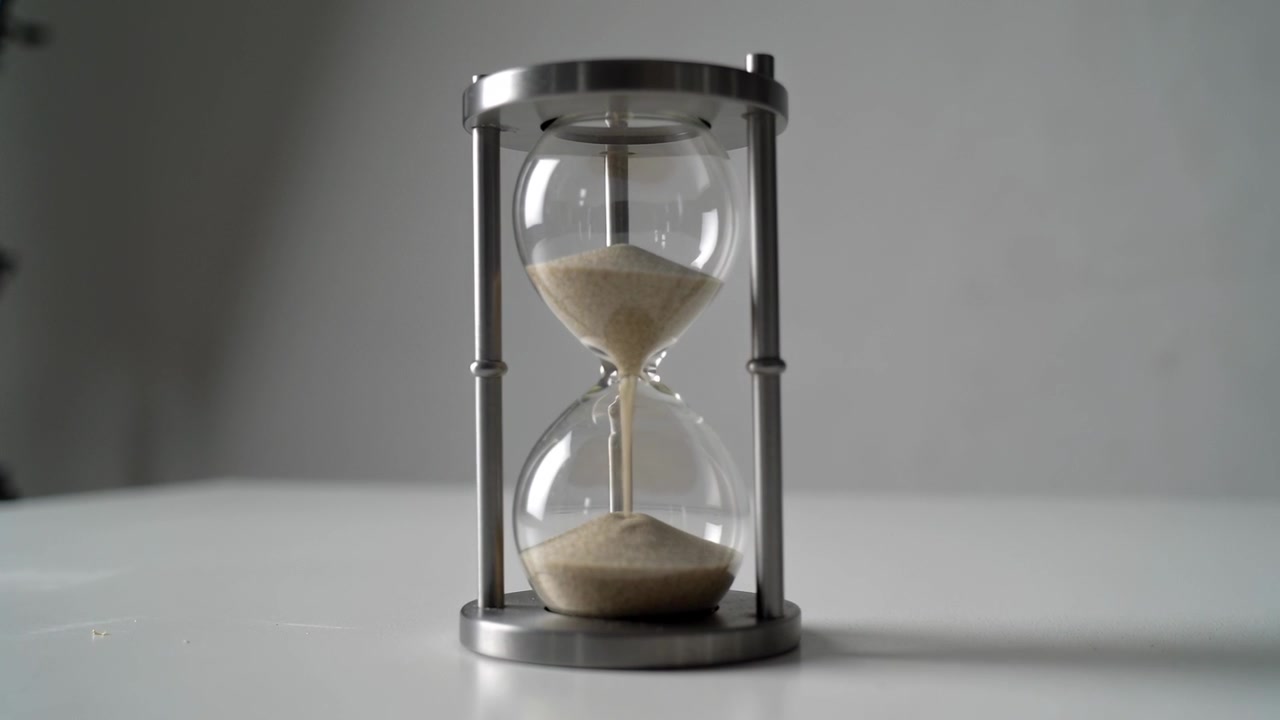 Gravity pulling beige sand, hourglass pouring sand through thin neck on white tabletop, metal frame