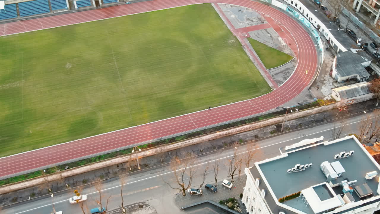 Aerial drone view of a stadium in Chisinau. Green field, running tracks with a man, tribunes, buildings nearby, roads with moving cars and bare trees. Moldova