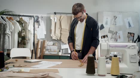 young male designer in eyeglasses in the workshop measure pieces of clothes in studio and smiles at camera