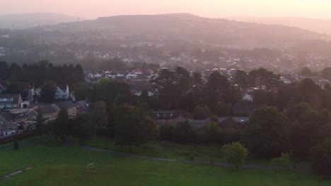 parallax moving aerial view of trees with houses on hillside at sunrise with recreational park in foreground. drone view of densely populated estates and streets