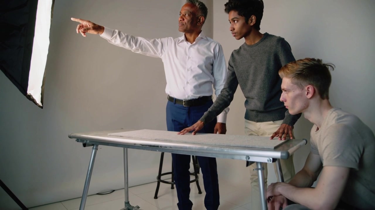 Ældre mand i hvid skjorte peger og vejleder to mænd, der læner sig over et bord i et studie, med softbox.