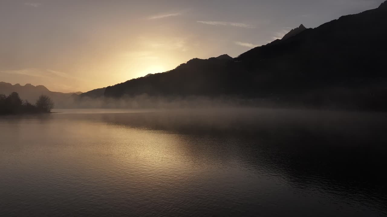 안개가 가득한 호수 위에서 아름다운 일출을 포착하는 정적 이미지, 오렌지색 빛이 안개를 고 물 위에 부드럽게 반사되어 조용하고 따뜻한 분위기를 만니다.