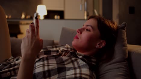 una chica morena triste e irritada con una camisa a cuadros se acuesta en el sofá escribiendo en redes sociales usando su teléfono inteligente blanco lo pone a un lado y mira irritadamente a un lado en un apartamento moderno por la noche