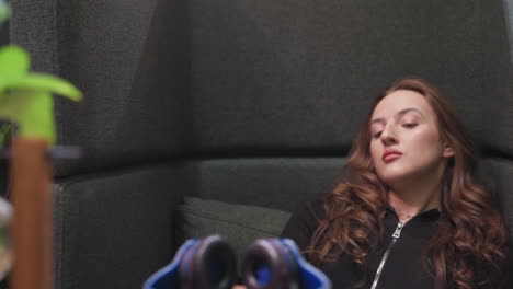 close up view of lady with long curly hair removing headset while resting on cushioned chair, head tilted upward with calm expression, creating serene atmosphere with soft focus