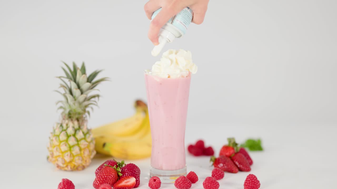 Hand tops creamy pink smoothie with whipped cream, surrounded by fresh fruit on white background