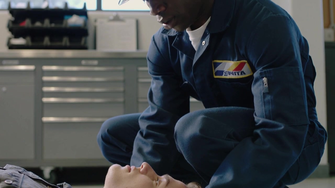 Un tecnico in tuta blu scuro stabilizza e sorregge la testa di un collega dopo il suo collasso, controllando le vie respiratorie in officina.