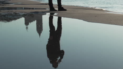 reflection-of-man-talking-on-a-phone,-then-strolling-along-coastline-as-sea-gently-washes-ashore-under-clear-sky-on-a-sunny-morning