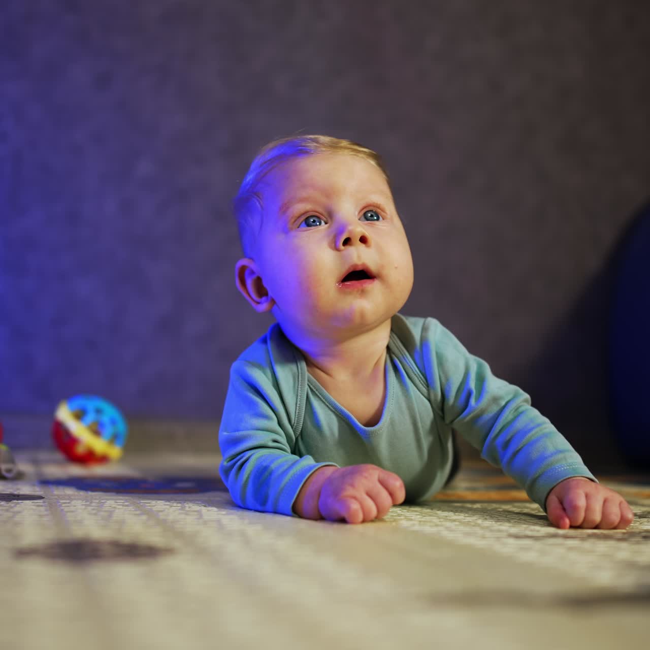 Bebê loiro adorável deitado de bruços olhando para cima com curiosidade. Criança fofa deitada no chão de casa em meio aos brinquedos