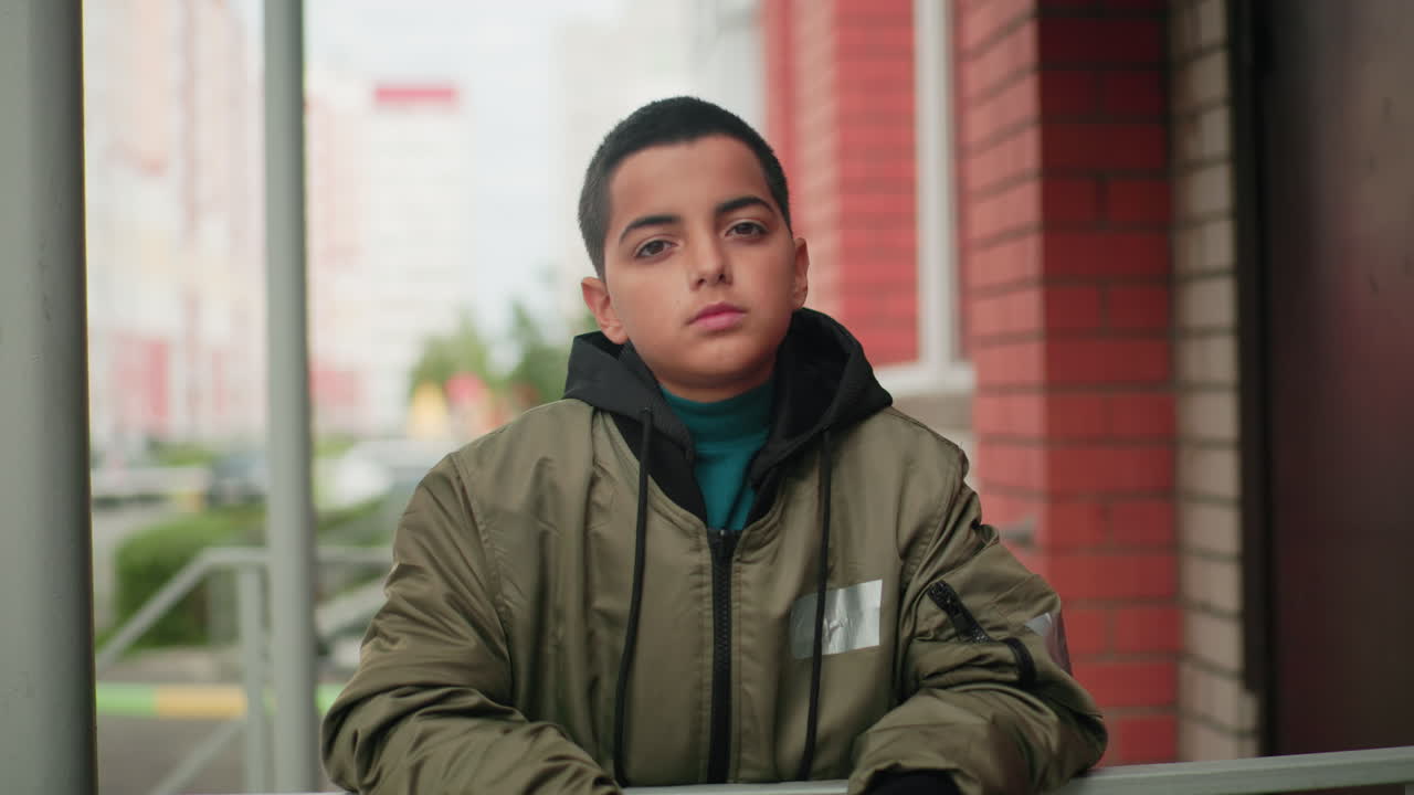 Un chico con una chaqueta verde está de pie delante de un edificio de ladrillo sujetando un libro cerrado y mirando hacia delante con expresión reflexiva. El fondo urbano, ligeramente desenfocado y con coches aparcados, crea una atmósfera pensativa.