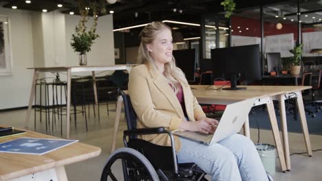 happy caucasian casual businesswoman in wheelchair using laptop in office, in slow motion