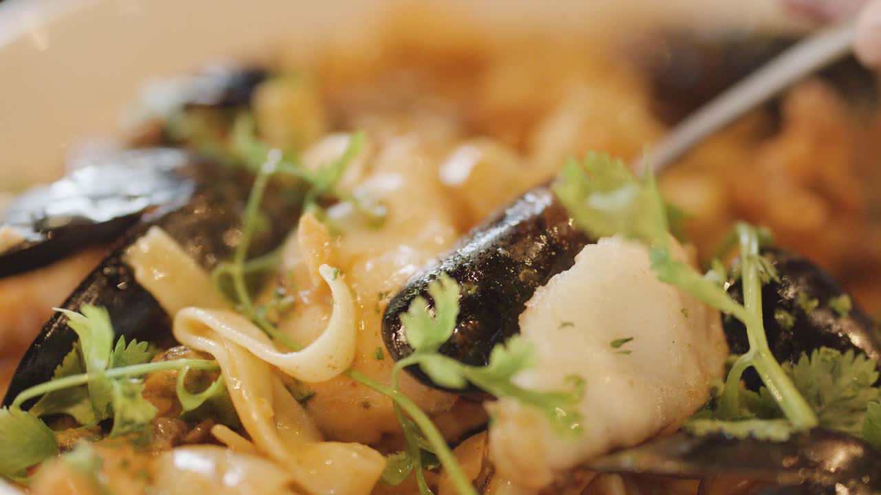 Macro view of assorted seafood pasta in a creamy sauce, garnished with herbs, as a hand uses a spoon to serve or eat