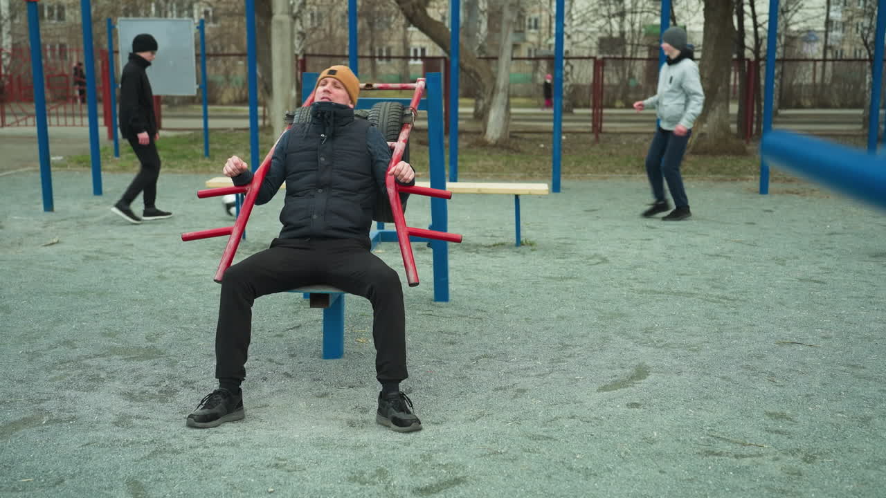 A coach in a brown beanie working out on an outdoor, with two boys in the background playing football, with blue pull up poles around